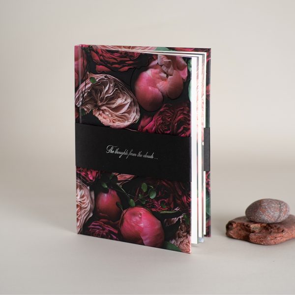 Pastatyta užrašinė, šalia du akmenukai, užrašinė apjuosta nuimama juosta, viršelis dekoruotas gėlių spauda